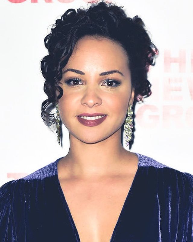Jasmine Cephas Jones smiling in a dark blue V-neck dress and silver earrings.